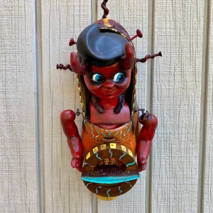 May include: A whimsical, handmade wall sculpture featuring a red devil-like figure with blue eyes, horns, and a black hat. The sculpture has outstretched arms and is mounted on a wooden background. The sculpture is made of metal and wood.