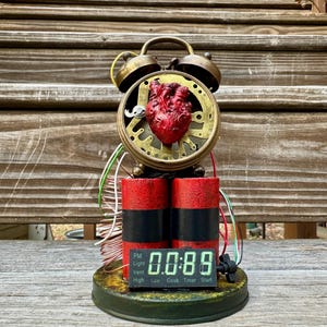 May include: A steampunk-style alarm clock with a red heart in the center. The clock features a digital display showing "00:89" and two red and black cylinders. The clock has a brass-colored bell and a green base.