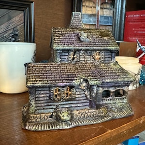 May include: A weathered, multi-level ceramic haunted house figurine. The house features a gray and purple color scheme with visible cracks and a chimney. The windows are lit, suggesting an interior light source. The figurine sits on a wooden shelf.