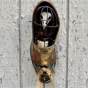 May include: A mixed-media sculpture featuring a bird skull encased in a metal cage, mounted on a shoe-shaped base. The sculpture incorporates brass, copper, and other metallic elements, with a distressed, aged aesthetic. The piece includes a small chain and a decorative fringe.