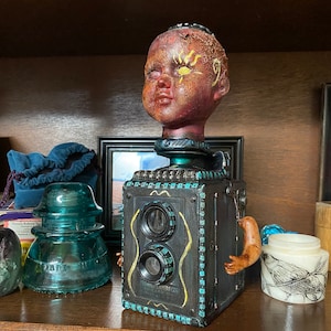 May include: A unique, handcrafted sculpture featuring a doll head atop a vintage camera body. The camera is black with teal and gold accents, and the doll head has a distressed, aged appearance. The sculpture includes a small arm attached to the side.