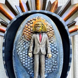 May include: A wall-mounted sculpture featuring a central figure in a tan suit and red tie, framed by a dark oval border with a sunburst-style top. The figure stands against a textured, honeycomb-patterned background with gold and blue gradients.