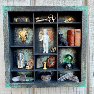 May include: A shadow box with a dark teal and black finish, containing various small objects. Items include a key, a skull, a small statue, a black bird, books, a ring, a glass ball, and crystals. The box is displayed against a wooden background.