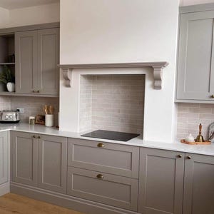 May include: A contemporary kitchen with grey cabinets, white worktops, and a light brick-effect splashback. The kitchen includes a built-in hob, brass handles, and wooden flooring. The design is clean and modern.