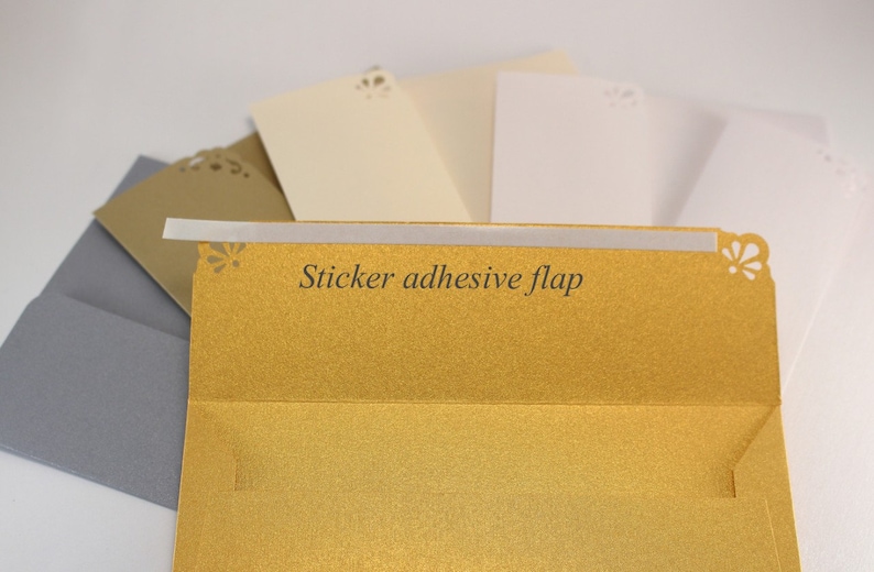 K&ouml;nnte beinhalten: Ein Satz Umschl&auml;ge in verschiedenen Farben, darunter Gold, Elfenbein und Grau. Der goldene Umschlag hat einen Klebeverschluss und ein dekoratives Ausschnittmuster.