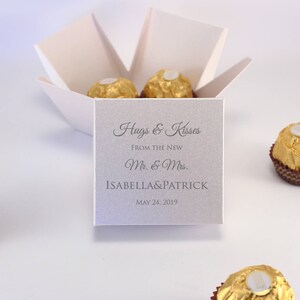 May include: A white favor box with a gray card insert that reads "Hugs & Kisses From the New Mr. & Mrs. ISABELLA&PATRICK MAY 24, 2019". The box is filled with chocolate candies wrapped in gold foil.