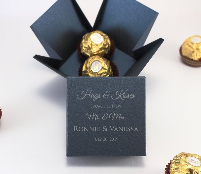 May include: A navy blue box with a lid that opens to reveal a small box with the text "Hugs & Kisses" and "From the New Mr. & Mrs. Ronnie & Vanessa July 20, 2019". The box is filled with chocolate candies.