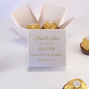 May include: White favor box with a gold inscription that reads "Hugs & Kisses From the New Mr. & Mrs. Jeannette & Robert September 3, 2019". The box contains four gold-wrapped chocolate candies.
