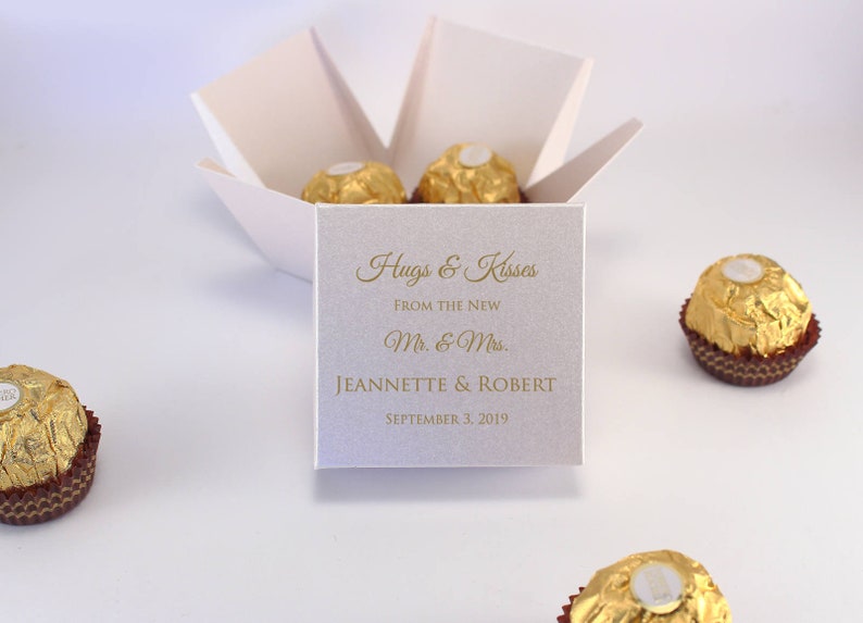 May include: White favor box with a gold inscription that reads "Hugs & Kisses From the New Mr. & Mrs. Jeannette & Robert September 3, 2019". The box contains four gold-wrapped chocolate candies.