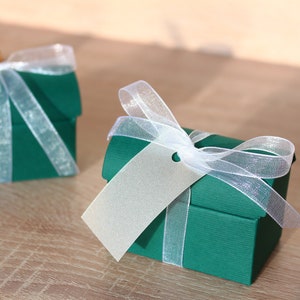 May include: Two small green gift boxes with white ribbon and a white tag. The boxes are tied with white ribbon and have a white tag attached to them. The boxes are on a wooden surface.