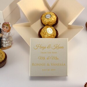 May include: A white box with a gold inscription that reads "Hugs & Kisses From the New Mr. & Mrs. Ronnie & Vanessa July 20, 2019". The box is filled with gold-wrapped chocolates.