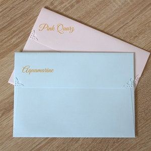 May include: Two envelopes, one light blue and one light pink, with decorative cutouts and the words "Aquamarine" and "Pink Quartz" printed in gold.