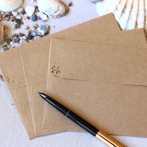 Puede incluir: Cuatro sobres de papel kraft marr&oacute;n con recortes decorativos en la solapa. Un bol&iacute;grafo negro y dorado est&aacute; sobre los sobres.