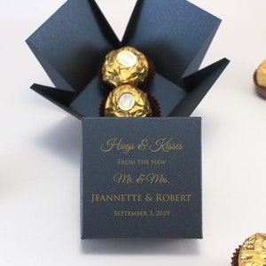 May include: A navy blue box with a gold inscription that reads "Hugs & Kisses From the New Mr. & Mrs. JEANNETTE & ROBERT SEPTEMBER 3, 2019". The box contains chocolate truffles wrapped in gold foil.