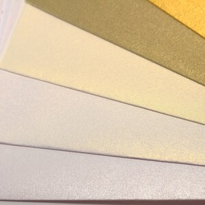 May include: A fan of different colored paper with a subtle shimmer. The colors are white, cream, light yellow, gold, and green.