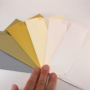 K&ouml;nnte beinhalten: Ein F&auml;cher aus sechs Umschl&auml;gen in verschiedenen Farben, darunter Silber, Gold und Elfenbein. Jeder Umschlag hat oben ein dekoratives Ausschnittmuster.