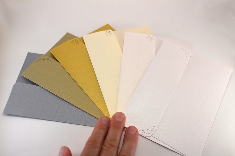 K&ouml;nnte beinhalten: Ein F&auml;cher aus sechs Umschl&auml;gen in verschiedenen Farben, darunter Silber, Gold und Elfenbein. Jeder Umschlag hat oben ein dekoratives Ausschnittmuster.