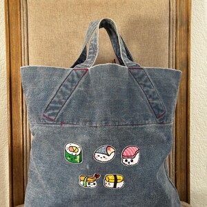 Può includere: Una borsa tote in denim con cuciture rosse e toppe a tema sushi. Le toppe raffigurano vari rotoli di sushi con facce di cartoni animati. La borsa ha due manici ed è appoggiata su una sedia. Misura circa 30 cm di larghezza e 35 cm di altezza.