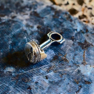 May include: A silver-colored metal charm in the shape of a car engine piston. The piston is detailed with a small, stylized wing design.