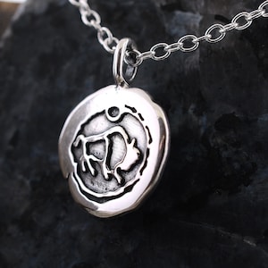 May include: A silver necklace with a round pendant featuring a stylized image of a bull. The pendant has a textured surface and a raised edge.