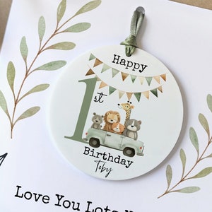 May include: A white card with green leafy vines on the sides. The card says "Happy 1st Birthday Toby Love You Lots Nanny" and has a removable round keepsake attached to it. The keepsake is green and white with a number 1, animals in a car, and the words "Happy Birthday".