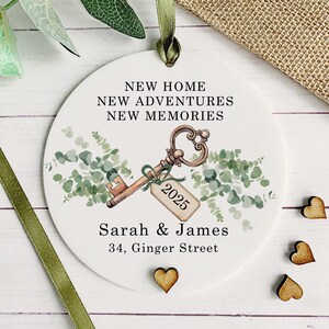 May include: White round ornament with a watercolor illustration of a key with a tag that says "2025" and greenery. The text "New Home, New Adventures, New Memories" is written above the key. Below the key is the text "Sarah & James, 34, Ginger Street".