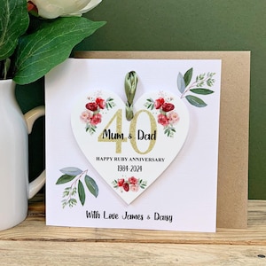 May include: A heart-shaped anniversary card with the text "Mum & Dad" and "40" in gold glitter. The card is decorated with red and pink floral accents and green leaves. The text "Happy Ruby Anniversary 1984-2024" is also included.