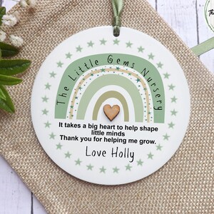 May include: A white round wooden sign with a green and beige rainbow design and the text "The Little Gems Nursery". The sign also includes the text "It takes a big heart to help shape little minds. Thank you for helping me grow. Love Holly."