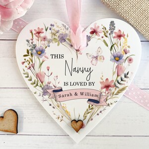 May include: A heart-shaped plaque with a floral design and the text "THIS Nanny IS LOVED BY Sarah & William". It has a pink ribbon for hanging. Two small wooden hearts are next to the plaque.