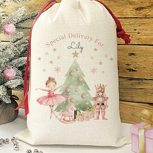 May include: A white canvas gift bag with a red drawstring. The bag features a watercolour illustration of a ballerina, a nutcracker, a rocking horse, and a Christmas tree. The text "Special Delivery For Lily" is printed on the bag.