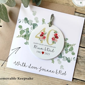 May include: A white round keepsake with gold glitter 40 and red floral accents. The text reads "Happy Ruby Anniversary Mum & Dad 1983 2023". The keepsake is attached to a white card with green foliage and the text "With Love Joanne & Rob Removable Keepsake".
