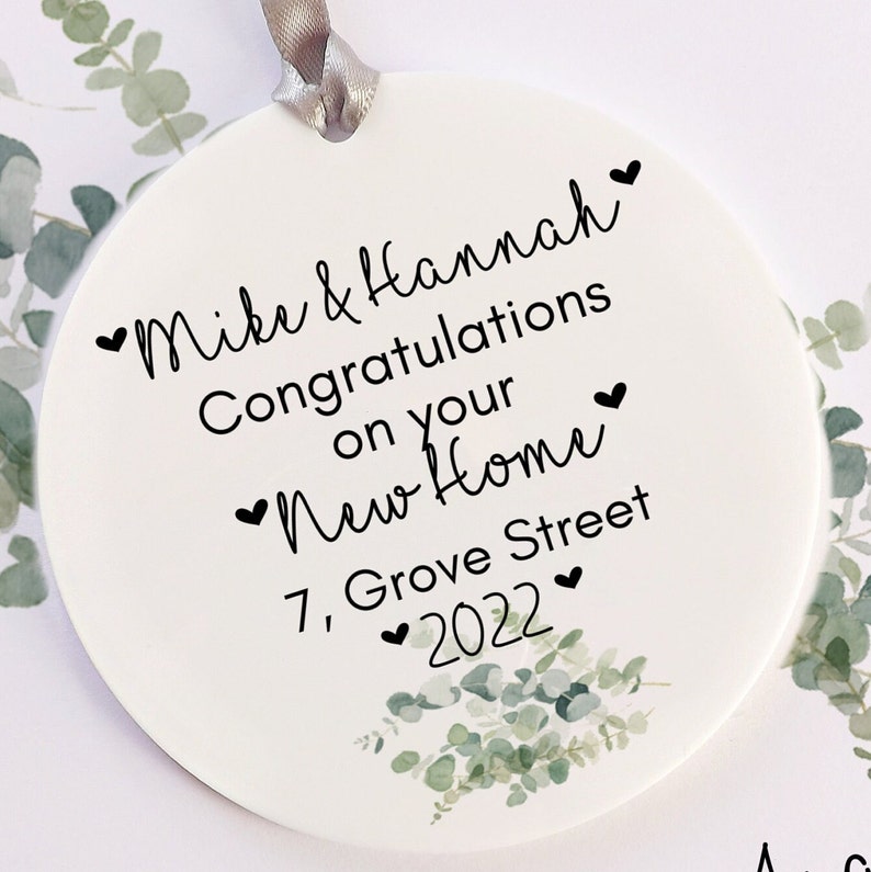Pu&ograve; includere: Ornamento rotondo bianco con una scritta nera che recita "Mike & Hannah Congratulazioni per la vostra nuova casa 7 Grove Street 2022" con piccoli cuori intorno al testo. L'ornamento &egrave; decorato con un disegno di foglie di eucalipto verdi.