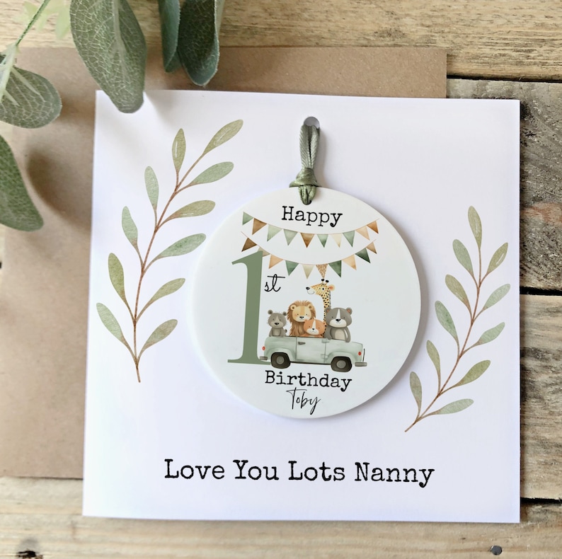 May include: A white card with a green and brown leafy design. A round, white, paper ornament is attached to the card with a green ribbon. The ornament has a green number one with the word "st" next to it. The ornament also has a picture of a green truck with a giraffe, lion, bear, and dog in the back. The ornament says "Happy Birthday Toby" at the bottom. The card says "Love You Lots Nanny" at the bottom.