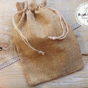 May include: A small, brown burlap drawstring bag with white drawstrings. The bag is sitting on a wooden surface with a white and gray ribbon with white polka dots.