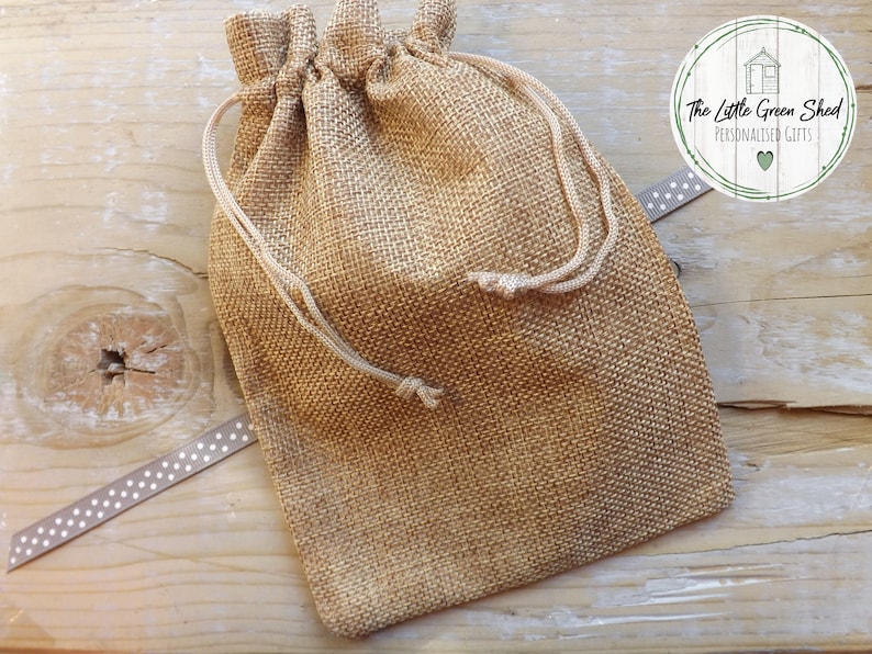 May include: A small, brown burlap drawstring bag with white drawstrings. The bag is sitting on a wooden surface with a white and gray ribbon with white polka dots.