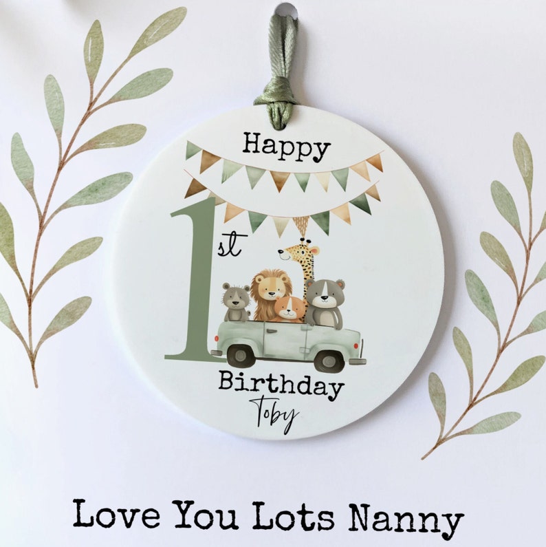 May include: A round, white, personalised ornament with a green number one, the words "Happy 1st Birthday Toby", and a green truck with animals. The truck has a giraffe, a lion, a dog, and two bears. The ornament has a green ribbon.