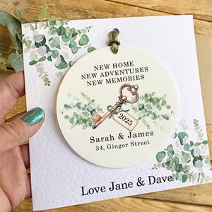 May include: A white round ornament with a green eucalyptus design and a gold key with a tag that says "2025". The ornament also has the text "New Home, New Adventures, New Memories", "Sarah & James", and "34, Ginger Street". The ornament is on a white card with the text "Love Jane & Dave".