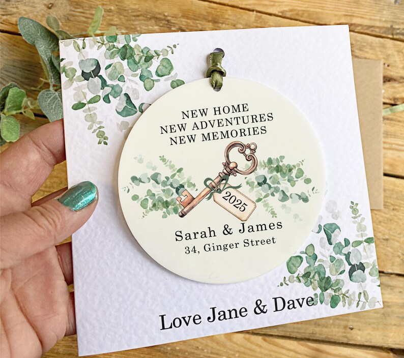 May include: A white round ornament with a green eucalyptus design and a gold key with a tag that says "2025". The ornament also has the text "New Home, New Adventures, New Memories", "Sarah & James", and "34, Ginger Street". The ornament is on a white card with the text "Love Jane & Dave".