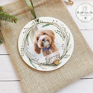 May include: A white ceramic Christmas ornament with a green wreath design, a photo of a brown dog, and the name "Barney" written in cursive. The ornament has a small wooden heart attached to it.