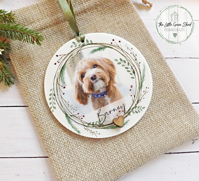 May include: A white ceramic Christmas ornament with a green wreath design, a photo of a brown dog, and the name "Barney" written in cursive. The ornament has a small wooden heart attached to it.