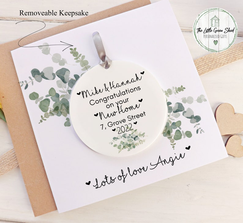 Pu&ograve; includere: Ciondolo in ceramica bianca con un design di eucalipto verde. Il ciondolo &egrave; personalizzato con il testo "Mike & Hannah Congratulazioni per la vostra nuova casa 7, Grove Street 2022" e "Tanto amore Angie".