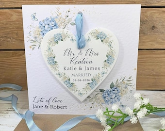 May include: A white square greeting card featuring a heart-shaped ornament with a blue ribbon. The card and ornament display blue hydrangea and butterfly illustrations. Text reads: Mr & Mrs Kentson, Katie & James, MARRIED 05-06-2026, and Lots of love, Jane & Robert.
