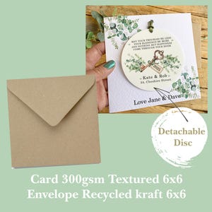 May include: A white card with a green floral design and a round, detachable disc with a key and the text "MAY YOUR TROUBLES BE LESS YOUR BLESSINGS BE MORE AND NOTHING BUT HAPPINESS COME THROUGH YOUR DOOR 2025 Kate & Rob 34. Cheshire Street Love Jane & Dave". The card is 6 inches by 6 inches and is made of 300gsm textured paper. The envelope is made of recycled kraft paper and is 6 inches by 6 inches.