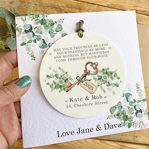 May include: A white round bauble with a green eucalyptus design and a gold key with the year 2025 on it. The bauble has the text "May your troubles be less, your blessings be more, and nothing but happiness come through your door."  The bauble also has the names "Kate & Rob" and the address "34, Cheshire Street". The bauble is attached to a card with the text "Love Jane & Dave".