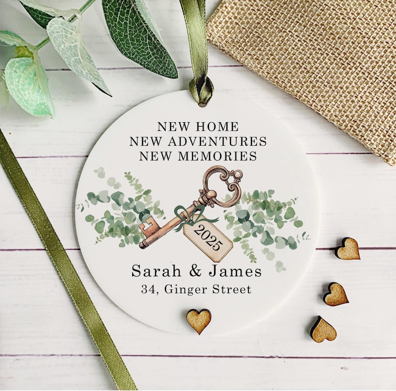 May include: White round ornament with a watercolor illustration of a key with a tag that says "2025" and greenery. The text "New Home, New Adventures, New Memories" is written above the key. Below the key is the text "Sarah & James, 34, Ginger Street".