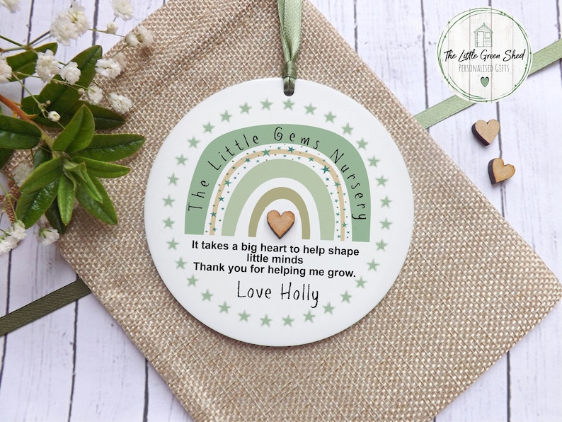 May include: A white round plaque with a green and beige rainbow design and the text "The Little Gems Nursery" and "It takes a big heart to help shape little minds. Thank you for helping me grow. Love Holly".