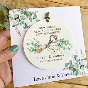 May include: A white round ornament with a green eucalyptus design and a gold key with a tag that says "2025". The ornament also has the text "New Home, New Adventures, New Memories", "Sarah & James", and "34, Ginger Street". The ornament is on a white card with the text "Love Jane & Dave".