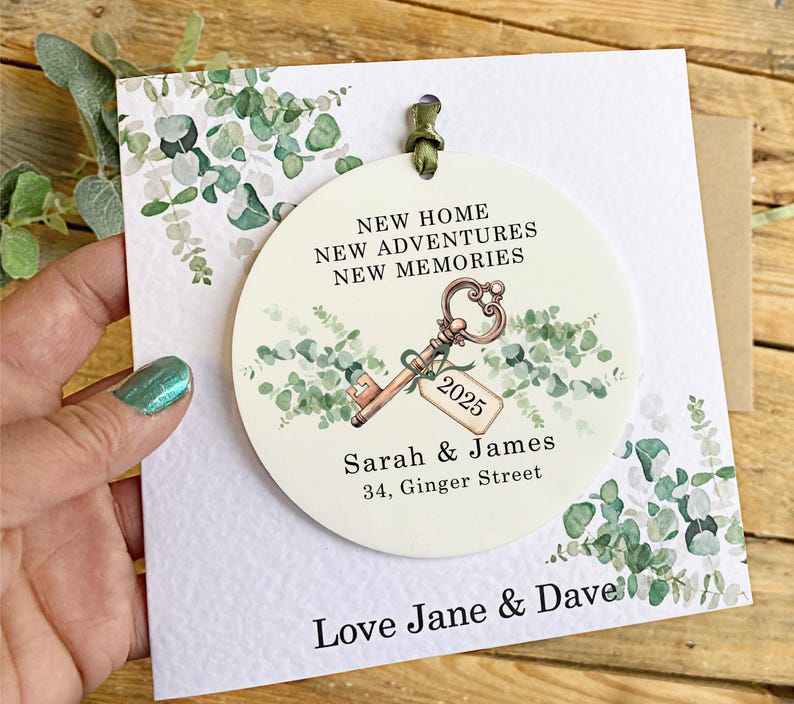 May include: A white round ornament with a green eucalyptus design and a gold key with a tag that says "2025". The ornament also has the text "New Home, New Adventures, New Memories", "Sarah & James", and "34, Ginger Street". The ornament is on a white card with the text "Love Jane & Dave".