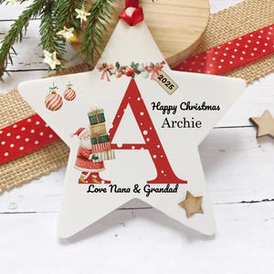 May include: A white star-shaped Christmas ornament with a large red letter "A" and the text "Happy Christmas Archie". The ornament features a Santa Claus illustration and the text "Love Nana & Grandad".