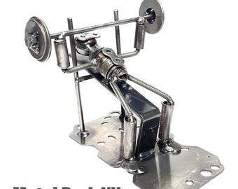 May include: A handcrafted metal sculpture of a weightlifter on a bench, made from various metal components. The sculpture features a barbell with weights, a bench, and a base. The text "Metal Park NY" is at the bottom.